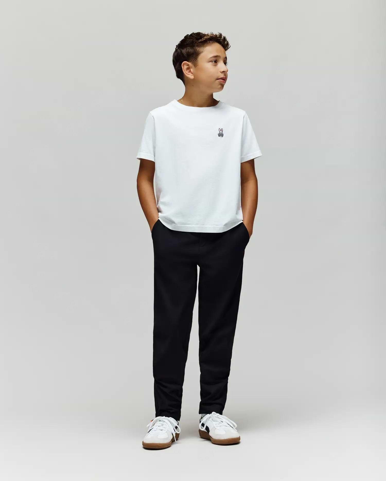 A boy stands against a plain light gray background, wearing a white t-shirt, Psycho Bunny KIDS MATHIEU COMMUTER PANT (KB0600002), and white sneakers. His hands are in his pockets, looking to the side.