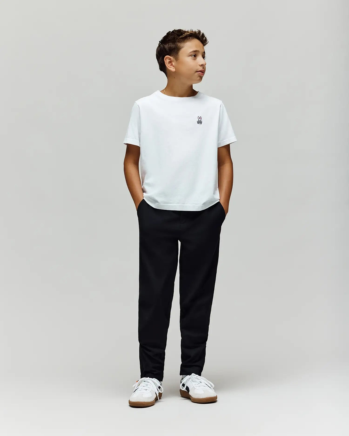 A boy stands against a plain light gray background, wearing a white t-shirt, Psycho Bunny KIDS MATHIEU COMMUTER PANT (KB0600002), and white sneakers. His hands are in his pockets, looking to the side.