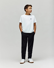 A boy stands against a plain light gray background, wearing a white t-shirt, Psycho Bunny KIDS MATHIEU COMMUTER PANT (KB0600002), and white sneakers. His hands are in his pockets, looking to the side.