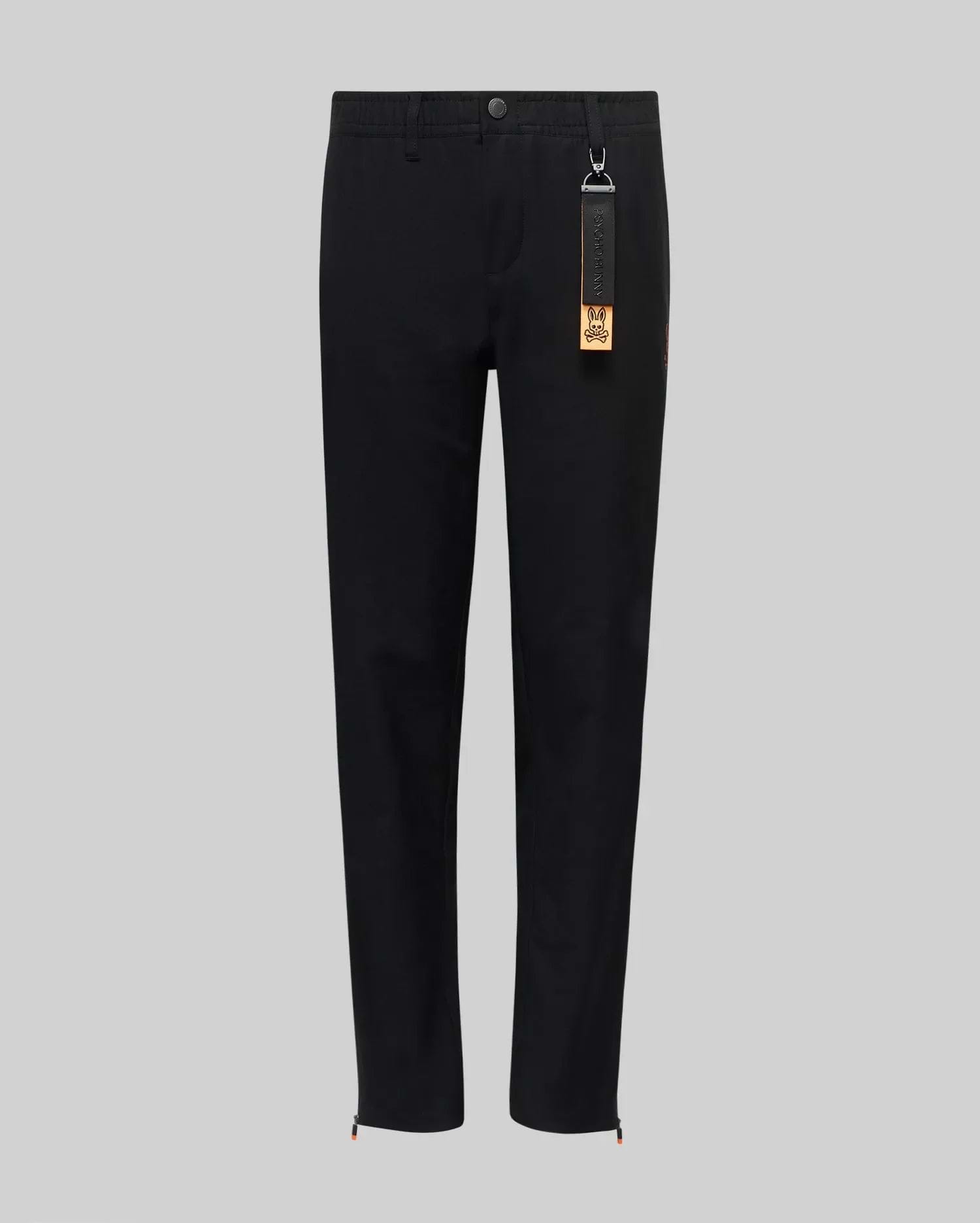The Psycho Bunny KIDS MATHIEU COMMUTER PANT (KB0600002) features a black straight-leg, classic fit with waistband, button and front zipper. A black tag with keychain and orange label hangs from a belt loop. Shown on a light gray background.