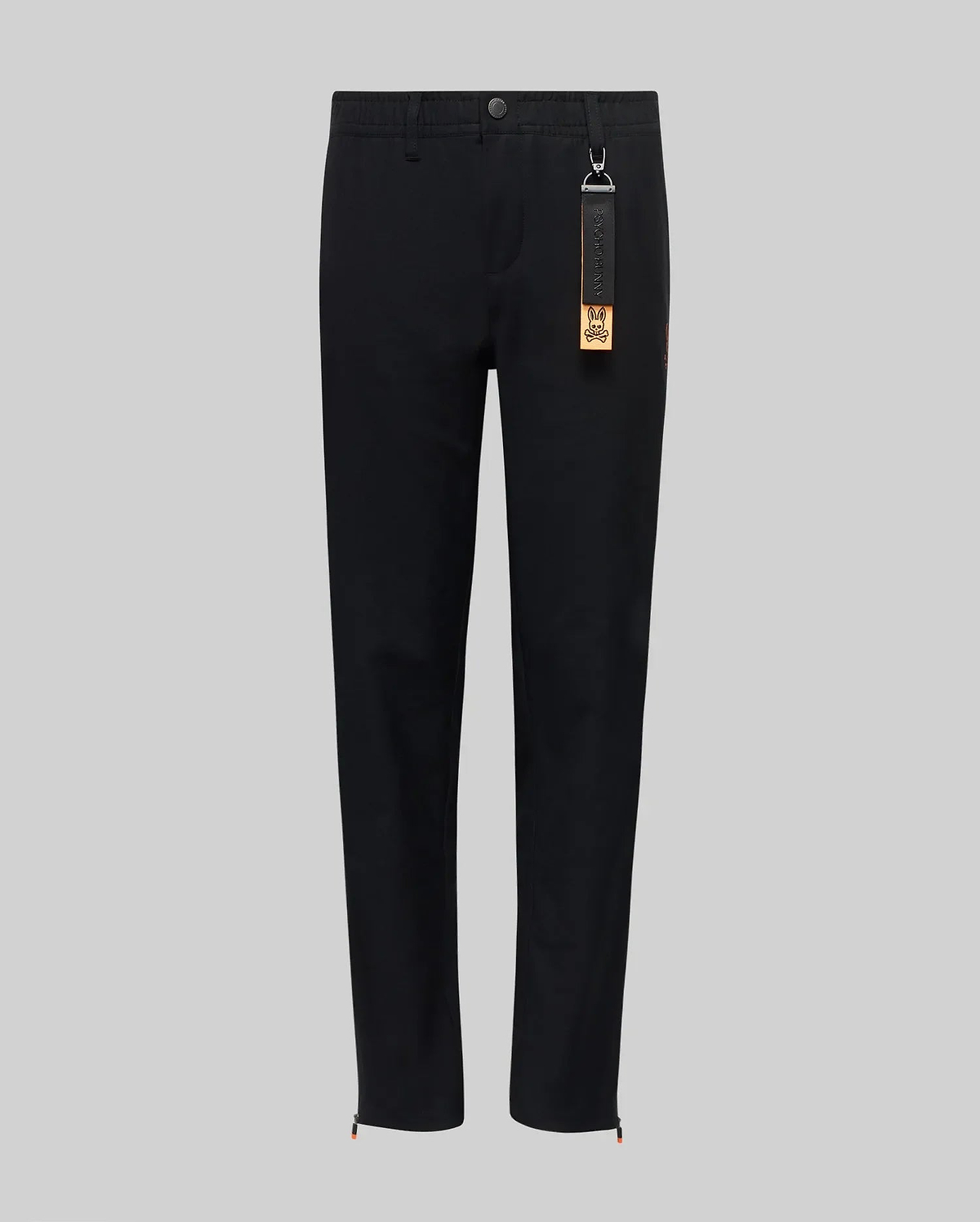 The Psycho Bunny KIDS MATHIEU COMMUTER PANT (KB0600002) features a black straight-leg, classic fit with waistband, button and front zipper. A black tag with keychain and orange label hangs from a belt loop. Shown on a light gray background.