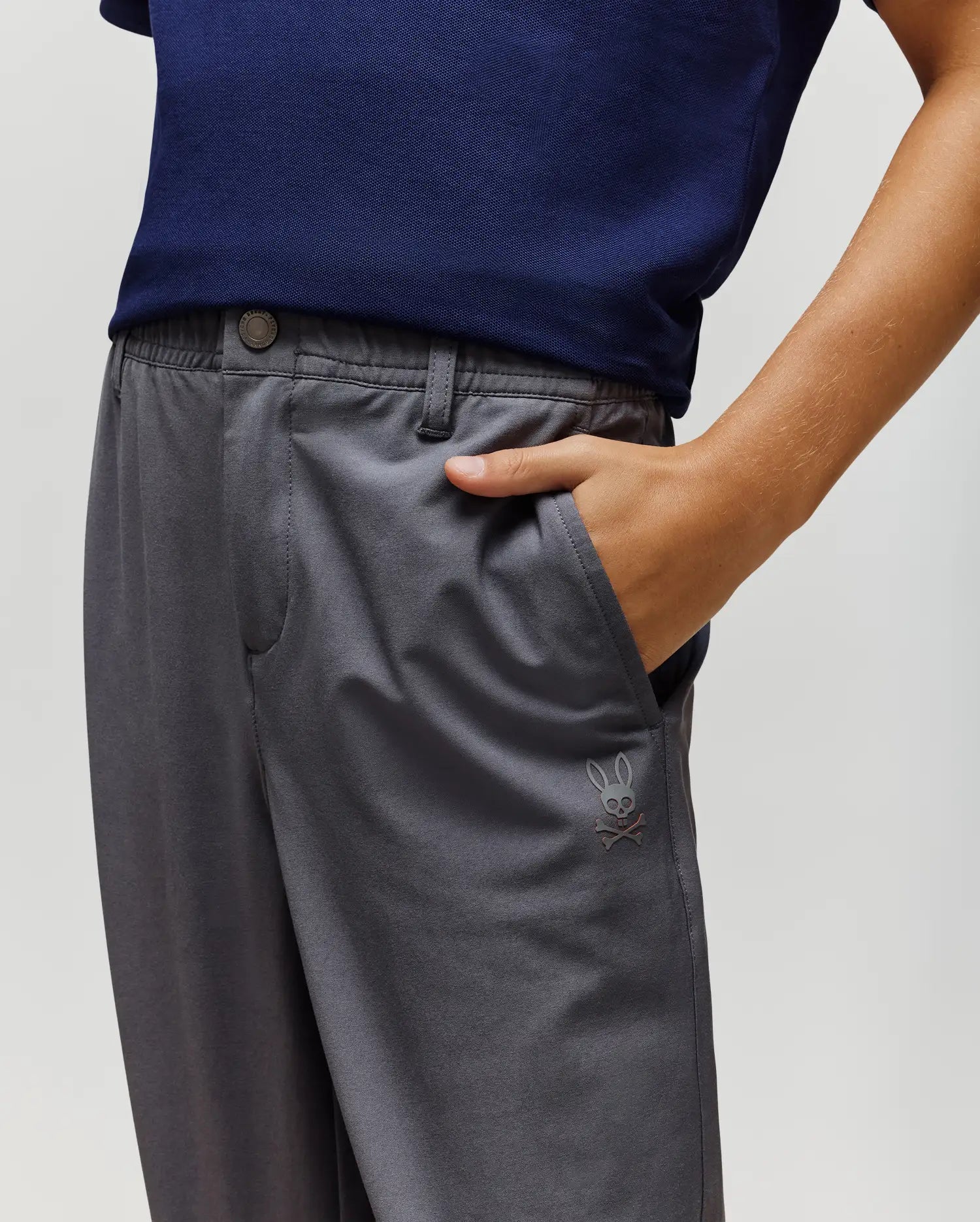 A person wears the Psycho Bunny KIDS MATHIEU COMMUTER PANT - KB0600002 in classic fit gray with a navy blue shirt; the pants feature a small Bunny rabbit skull and crossbones embroidery near the pocket.