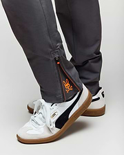 Close-up of a person wearing Psycho Bunny KIDS MATHIEU COMMUTER PANT - KB0600002 in gray with ankle zippers and orange graphic detail, paired with white Puma sneakers featuring black stripes and gum soles.