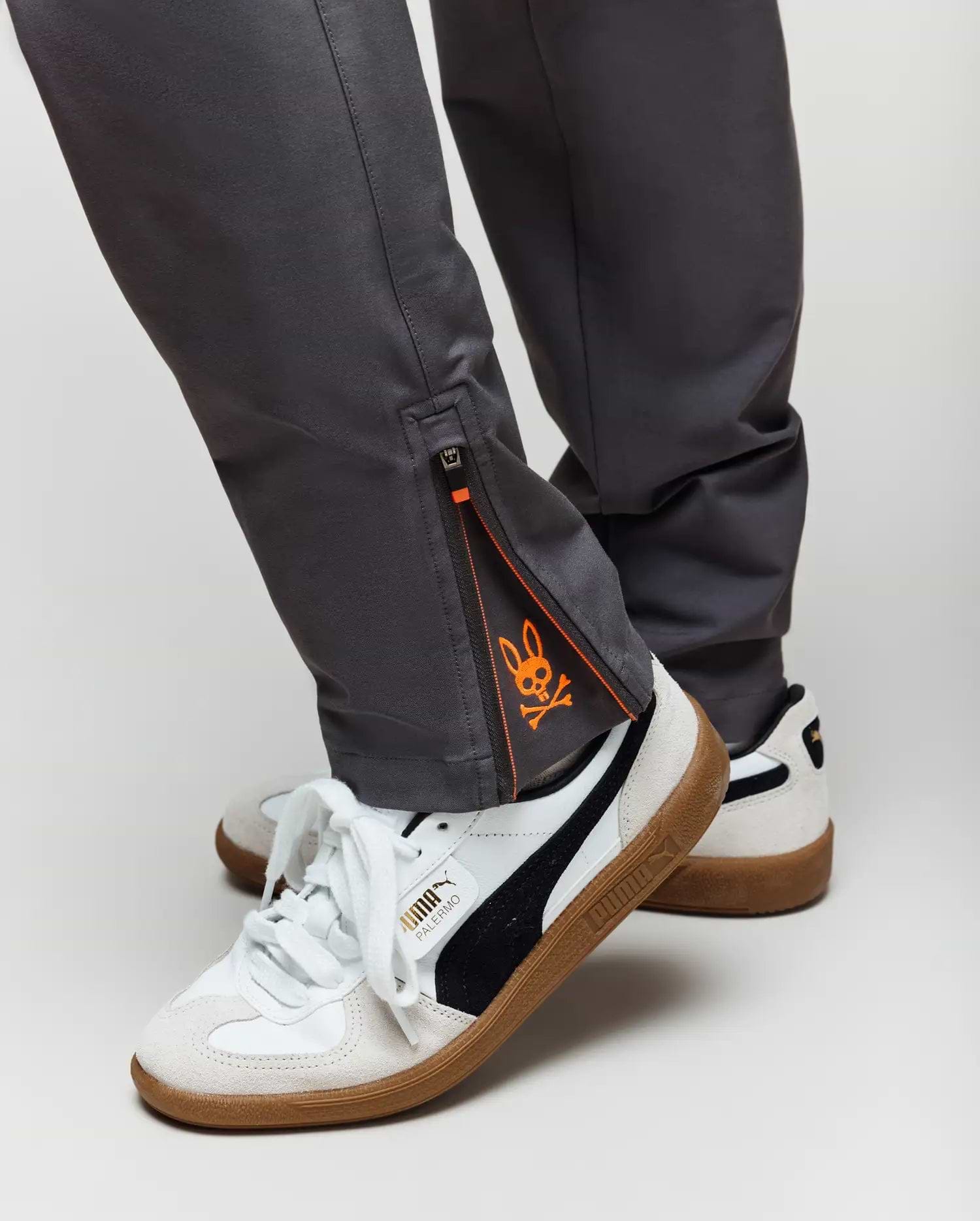 Close-up of a person wearing Psycho Bunny KIDS MATHIEU COMMUTER PANT - KB0600002 in gray with ankle zippers and orange graphic detail, paired with white Puma sneakers featuring black stripes and gum soles.