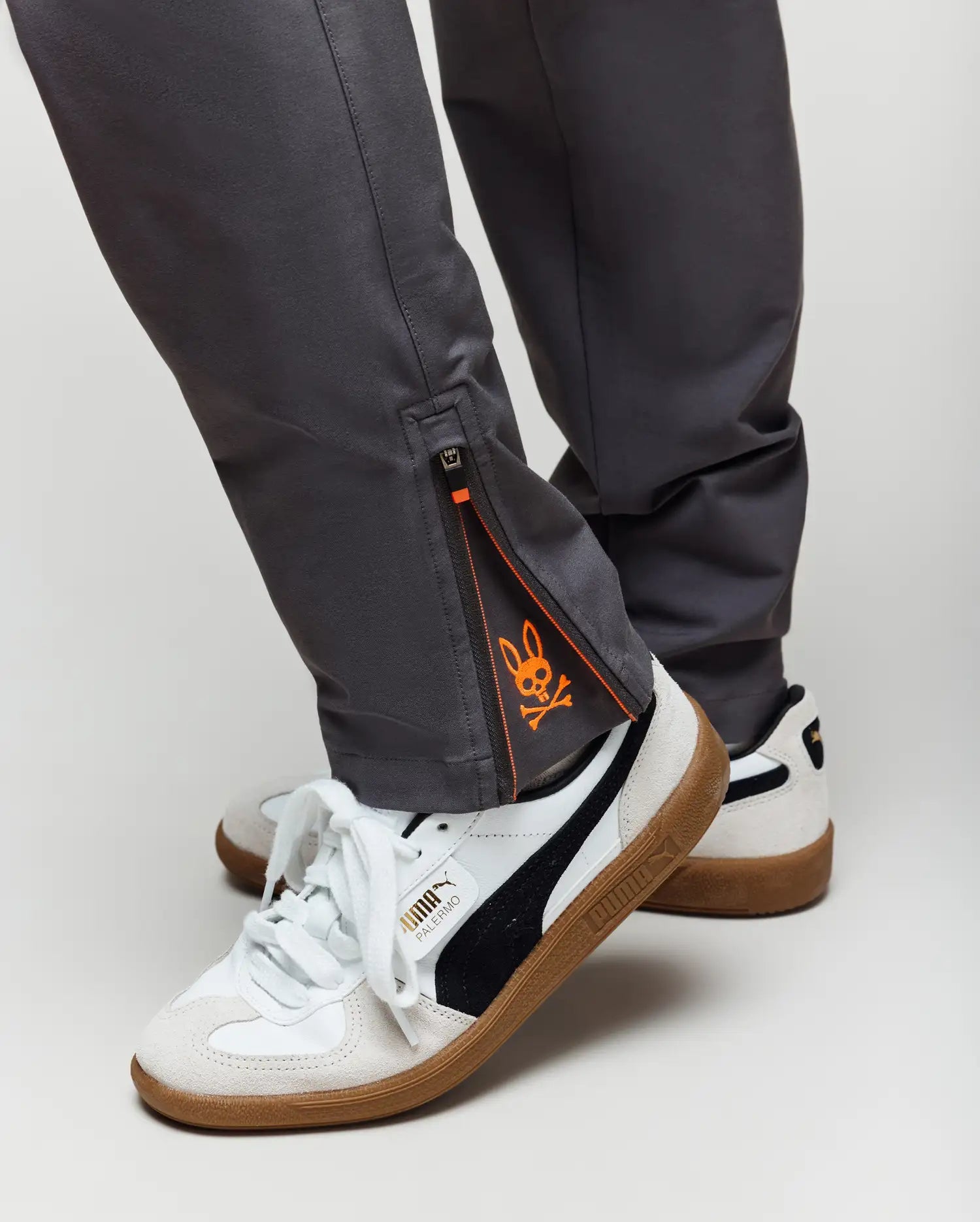 Close-up of a person wearing Psycho Bunny KIDS MATHIEU COMMUTER PANT - KB0600002 in gray with ankle zippers and orange graphic detail, paired with white Puma sneakers featuring black stripes and gum soles.