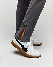 A person wears classic fit gray KIDS MATHIEU COMMUTER PANT by Psycho Bunny with orange and black "BUNNY OHOOHOH" zipper, Bunny embroidery, and white Puma sneakers featuring black stripes and gum soles.