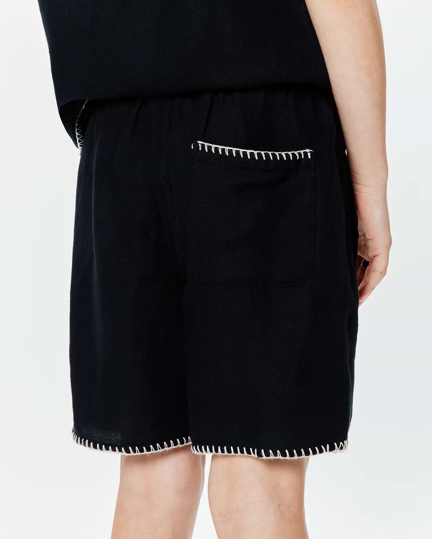 Kids Brad Linen Short in black, a relaxed fit garment with visible linen texture, accented by distinctive white blanket stitching on the back patch pocket and along the bottom hem.