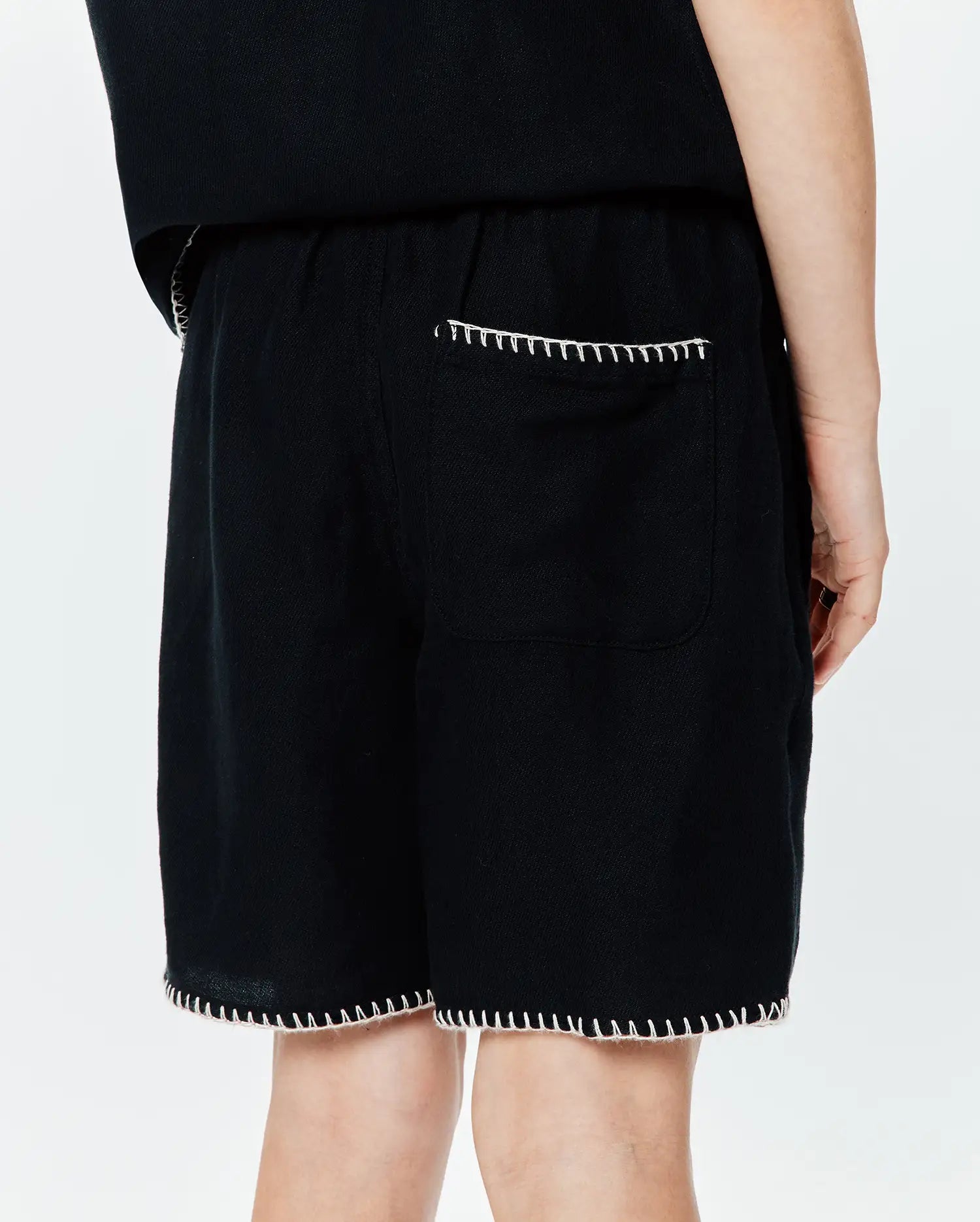 Kids Brad Linen Short in black, a relaxed fit garment with visible linen texture, accented by distinctive white blanket stitching on the back patch pocket and along the bottom hem.