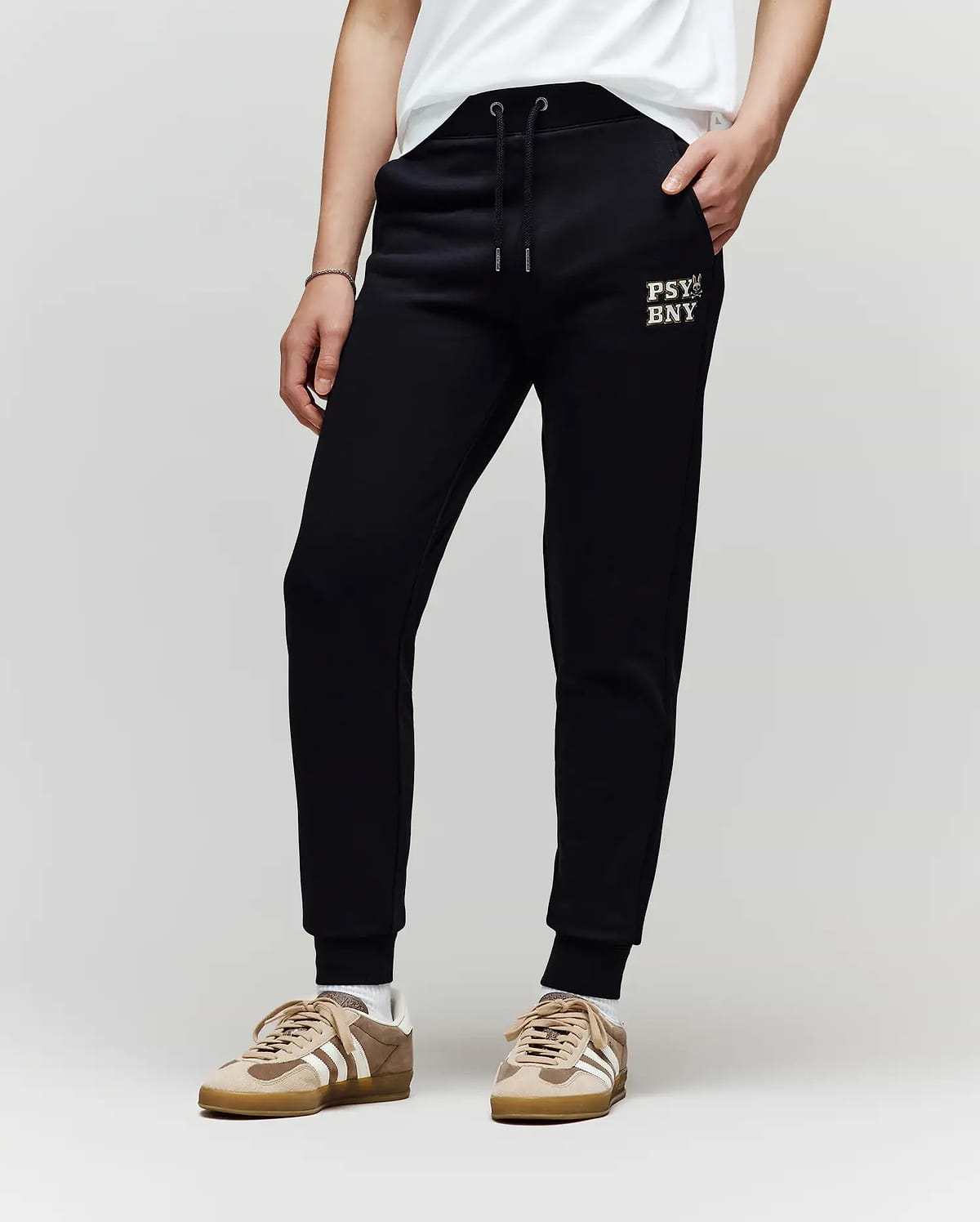 A child models the Psycho Bunny KIDS WADE CORD PATCH SWEATPANT (KB0800002), featuring an embroidered wordmark on the left thigh, paired with a white shirt and beige sneakers against a plain light background for a classic athletic look.