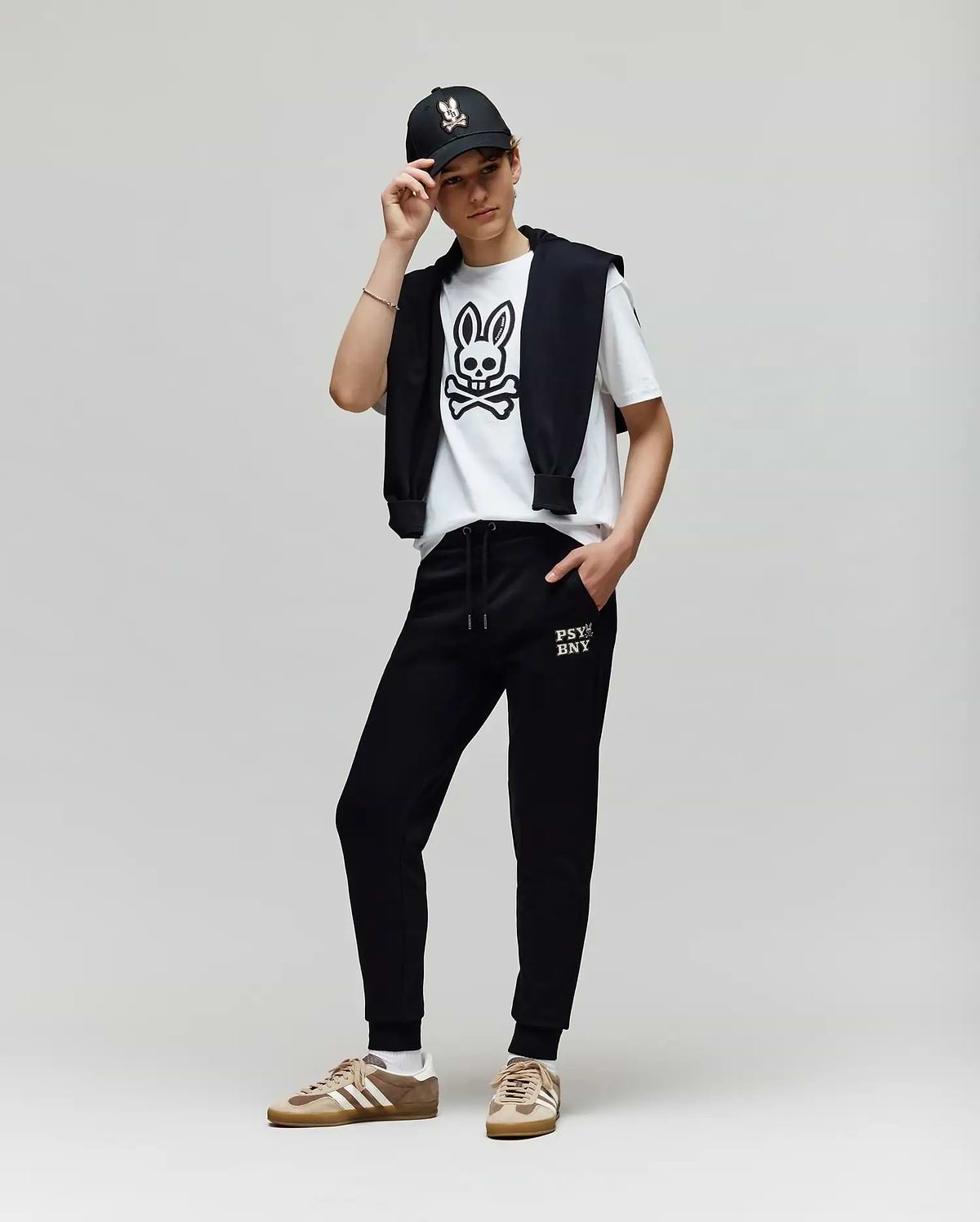 A person poses against a plain background, wearing a black cap, a white Psycho Bunny T-shirt with a bold cartoon bunny graphic, KIDS WADE CORD PATCH SWEATPANT - KB0800002 by Psycho Bunny, and tan Adidas sneakers.
