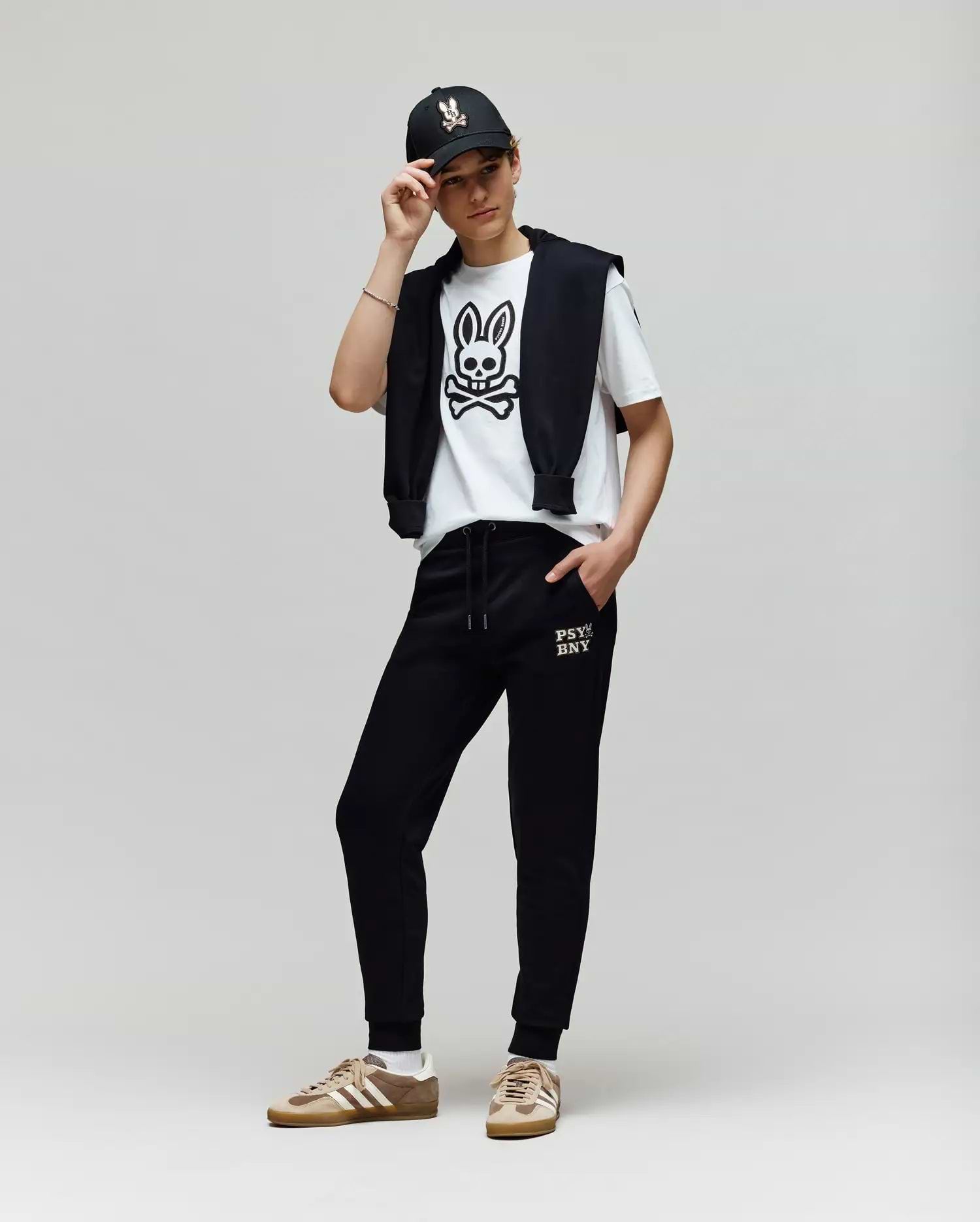 A person poses against a plain background, wearing a black cap, a white Psycho Bunny T-shirt with a bold cartoon bunny graphic, KIDS WADE CORD PATCH SWEATPANT - KB0800002 by Psycho Bunny, and tan Adidas sneakers.