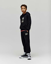 A boy with short curly hair stands against a plain background wearing Psycho Bunny KIDS WADE CORD PATCH SWEATPANT (KB0800002), a black hoodie with a bunny graphic, and black high-top sneakers, hands in his pockets.