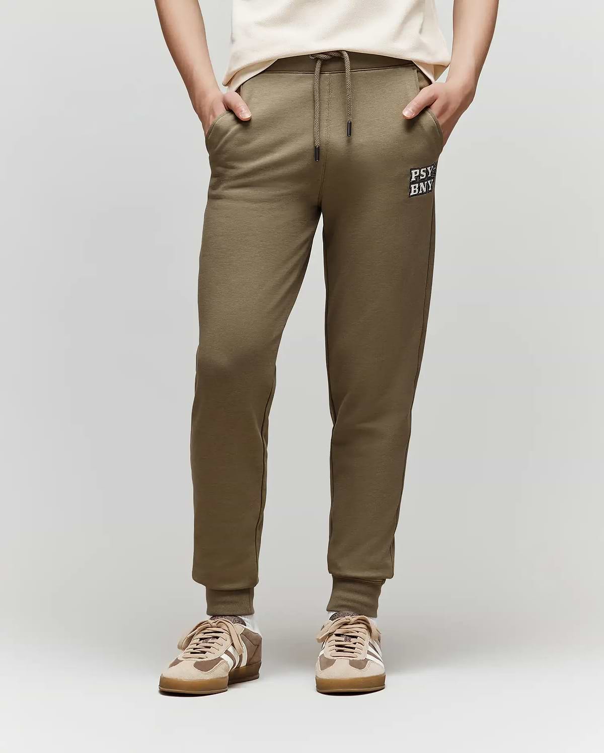 A child models the Psycho Bunny KIDS WADE CORD PATCH SWEATPANT (KB0800002) in olive green with a drawstring waist and "PSY BNK" text on the left thigh, paired with a white T-shirt and beige sneakers, standing against a plain background.