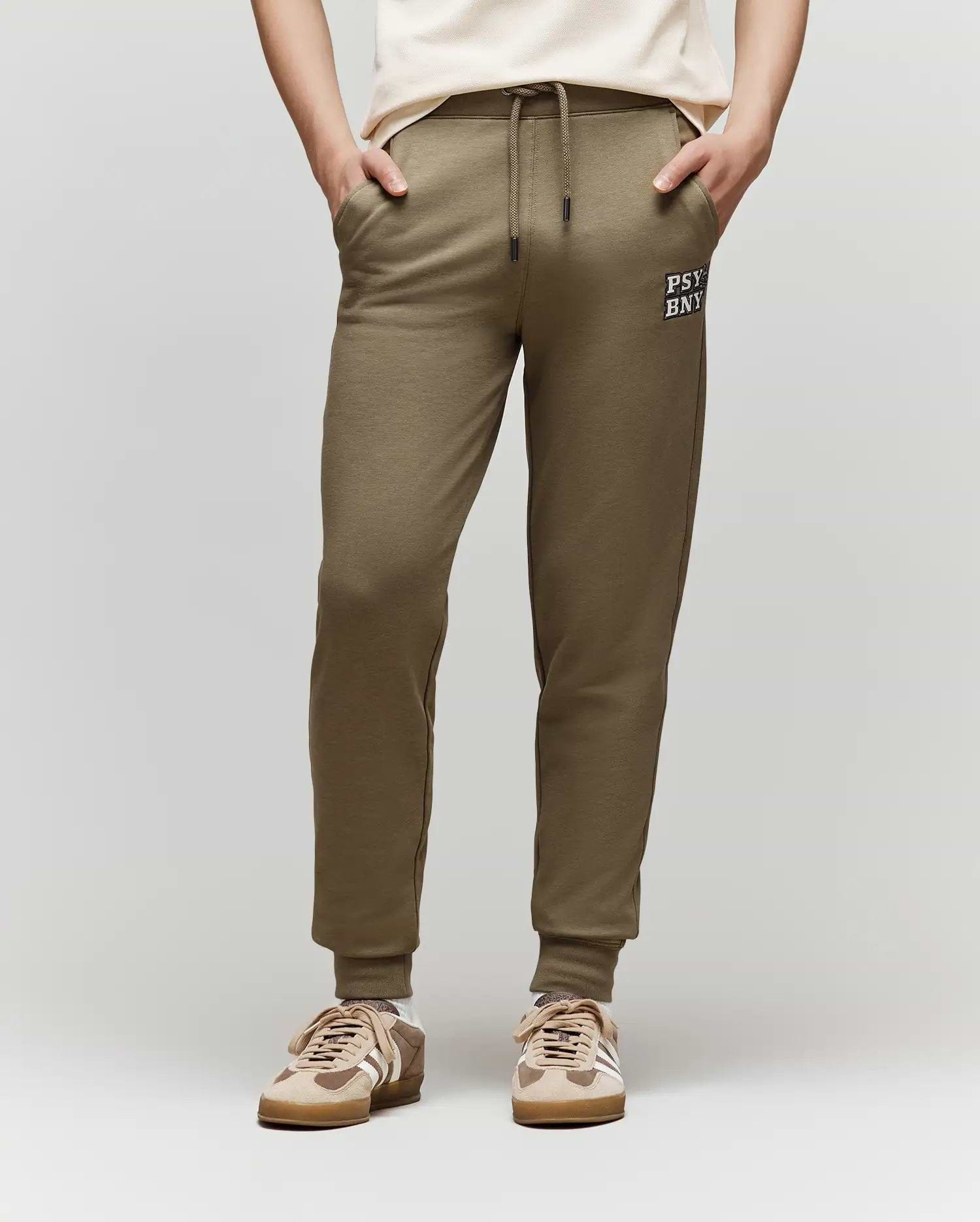 A child models the Psycho Bunny KIDS WADE CORD PATCH SWEATPANT (KB0800002) in olive green with a drawstring waist and "PSY BNK" text on the left thigh, paired with a white T-shirt and beige sneakers, standing against a plain background.