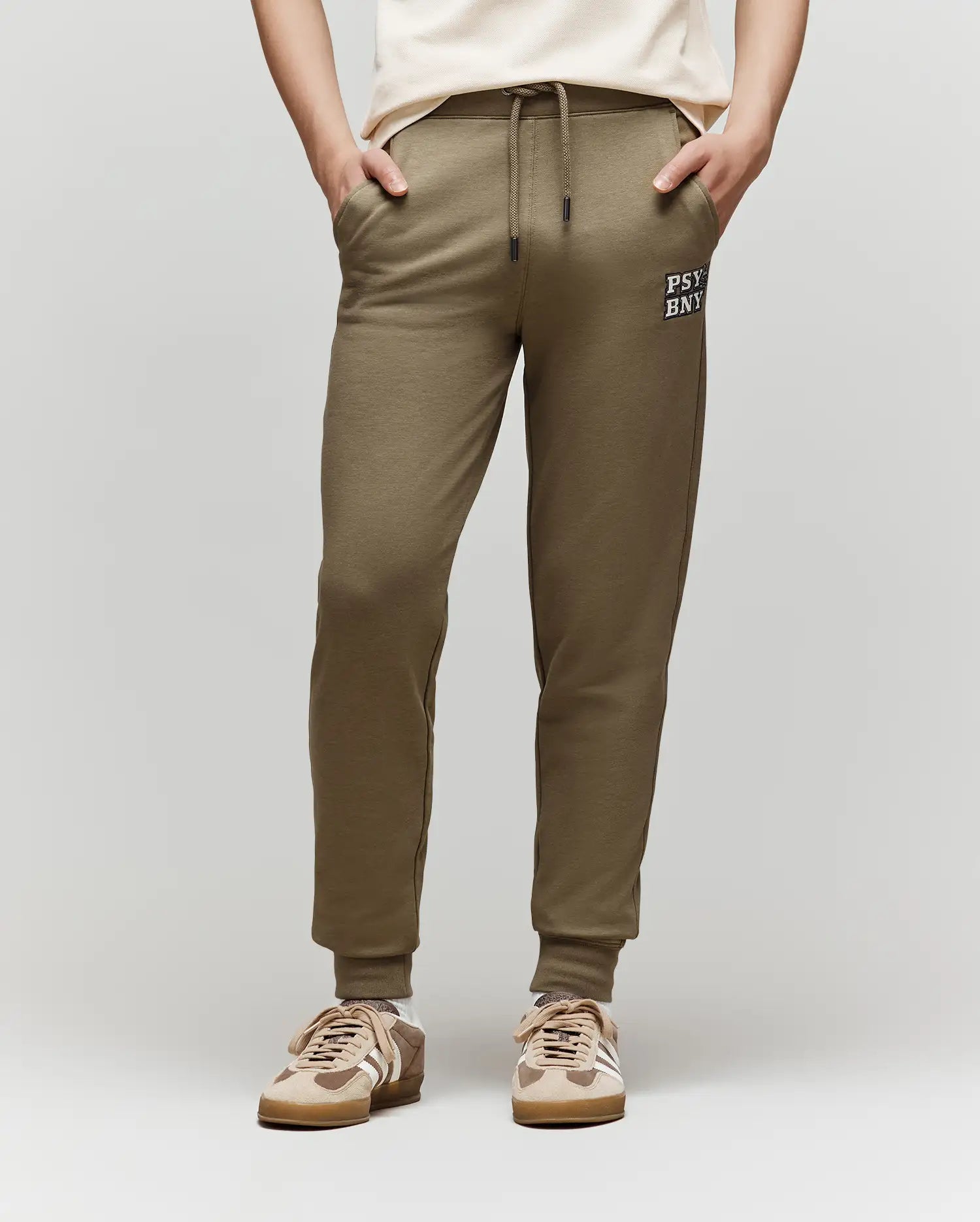 A child models the Psycho Bunny KIDS WADE CORD PATCH SWEATPANT (KB0800002) in olive green with a drawstring waist and "PSY BNK" text on the left thigh, paired with a white T-shirt and beige sneakers, standing against a plain background.