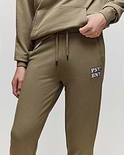 A person wears Psycho Bunny KIDS WADE CORD PATCH SWEATPANT (KB0800002), olive green joggers made from premium cotton blend, featuring a drawstring waist and “PSY BNYS” in white on the left thigh, with one hand in their pocket.