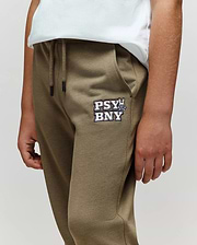 A person wears Psycho Bunny KIDS WADE CORD PATCH SWEATPANT (KB0800002) in olive green, featuring a drawstring waist, "PSY BNY" and cartoon bunny logo on the left pocket, paired with a white t-shirt. Only torso and upper legs are shown.