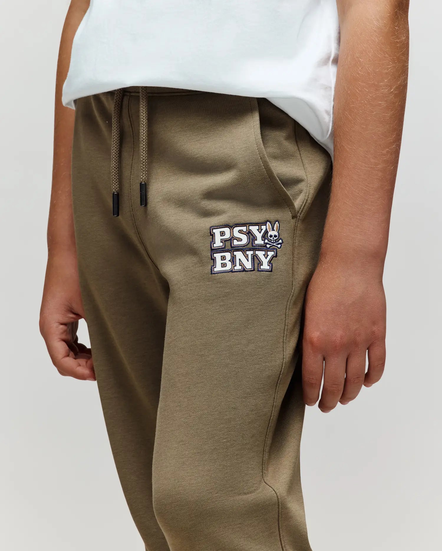 A person wears Psycho Bunny KIDS WADE CORD PATCH SWEATPANT (KB0800002) in olive green, featuring a drawstring waist, "PSY BNY" and cartoon bunny logo on the left pocket, paired with a white t-shirt. Only torso and upper legs are shown.