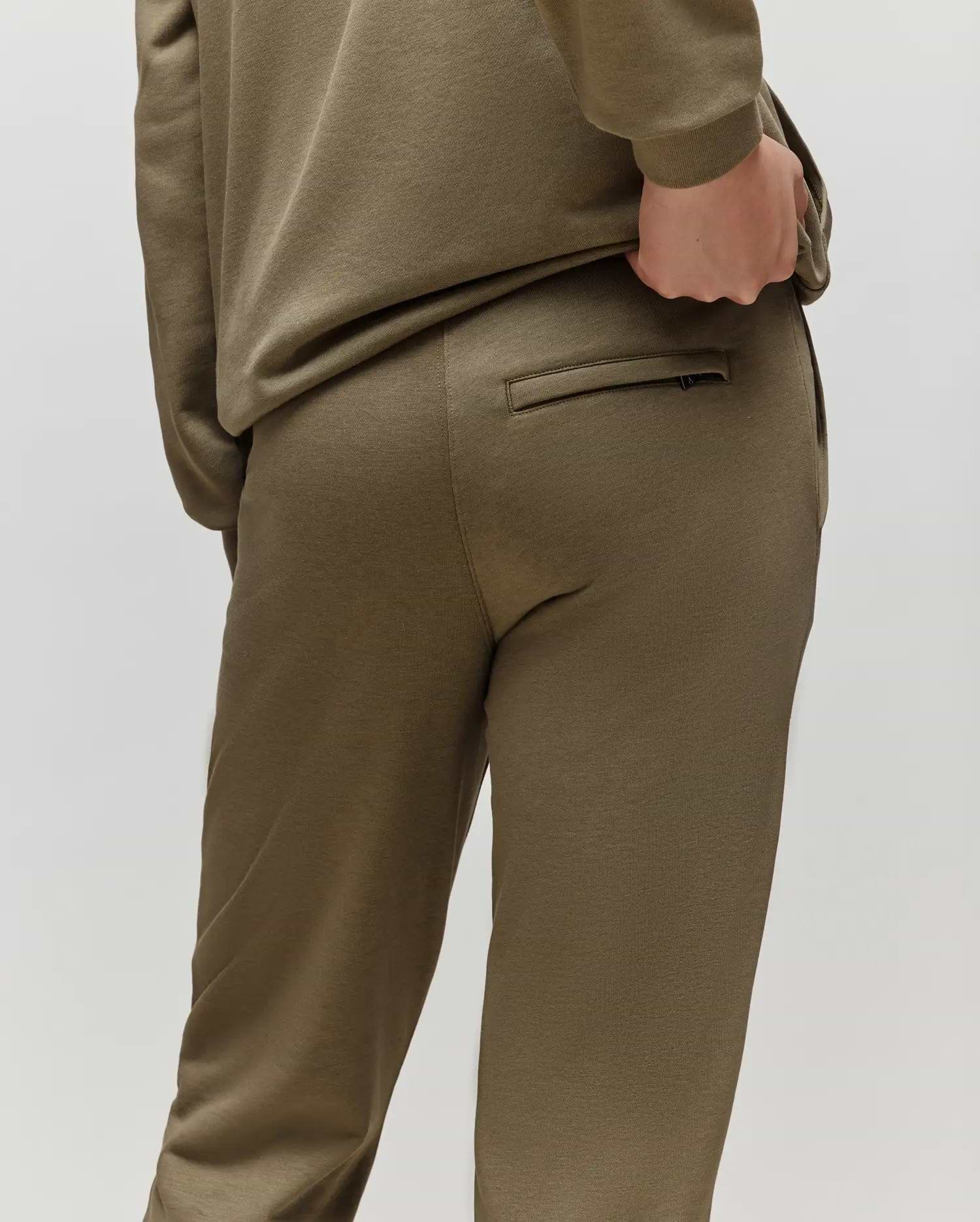 A child models Psycho Bunny's KIDS WADE CORD PATCH SWEATPANT (KB0800002) in olive green, featuring a drawstring waist, standing with a hand on their back pocket and facing away against a plain light background.