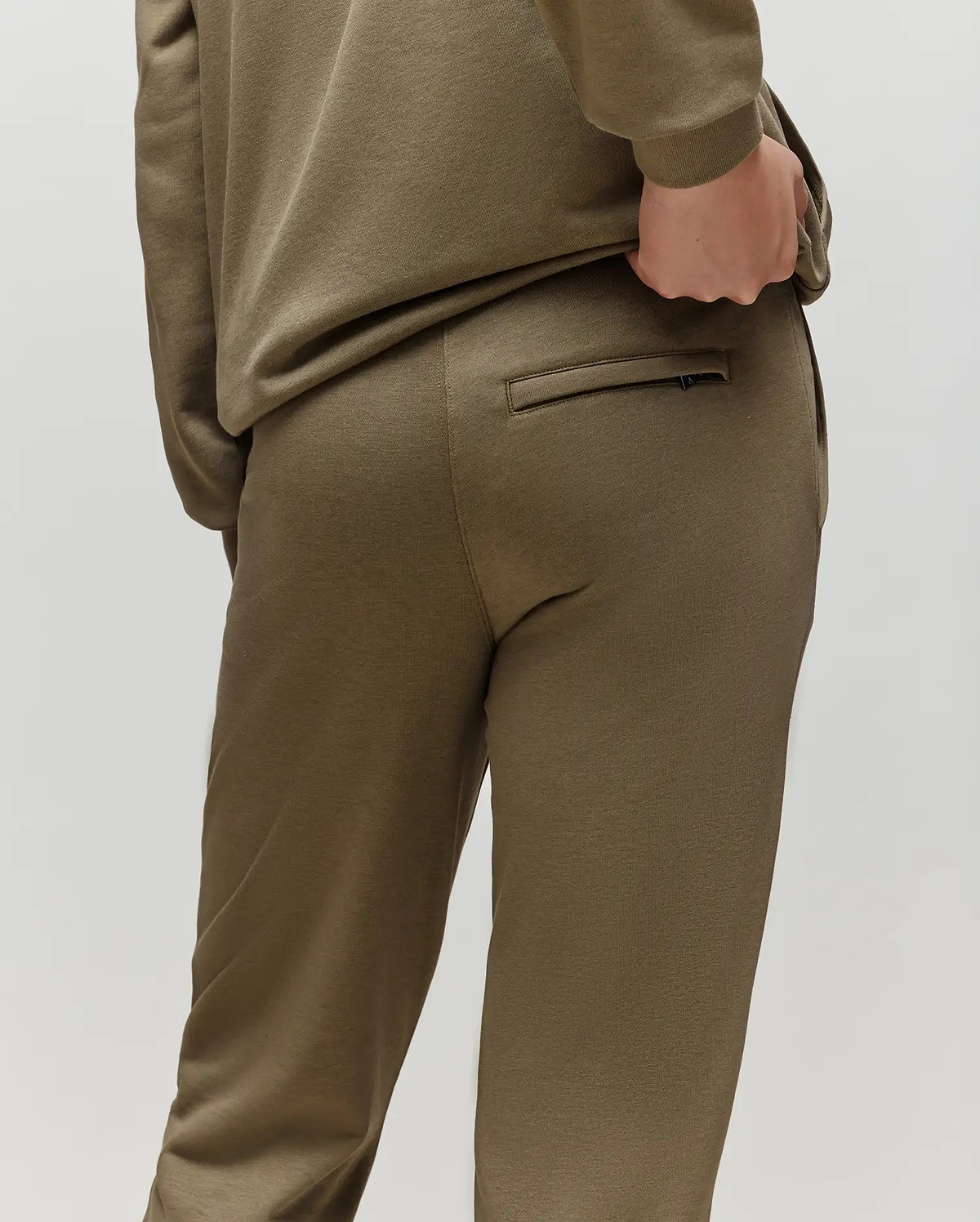 A child models Psycho Bunny's KIDS WADE CORD PATCH SWEATPANT (KB0800002) in olive green, featuring a drawstring waist, standing with a hand on their back pocket and facing away against a plain light background.