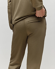 A child models Psycho Bunny's KIDS WADE CORD PATCH SWEATPANT (KB0800002) in olive green, featuring a drawstring waist, standing with a hand on their back pocket and facing away against a plain light background.