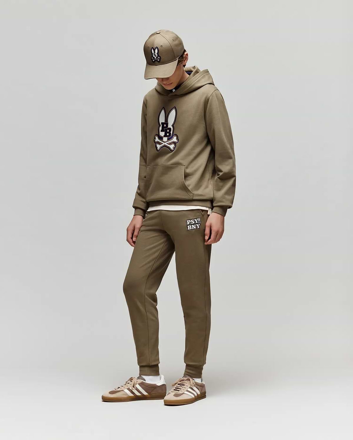 Dressed in Psycho Bunny’s KIDS WADE CORD PATCH SWEATPANT (KB0800002), a child stands against a plain light gray background, looking down with hands in pockets. They pair the brown sweatpants and hoodie with matching sneakers and a baseball cap.