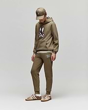 Dressed in Psycho Bunny’s KIDS WADE CORD PATCH SWEATPANT (KB0800002), a child stands against a plain light gray background, looking down with hands in pockets. They pair the brown sweatpants and hoodie with matching sneakers and a baseball cap.