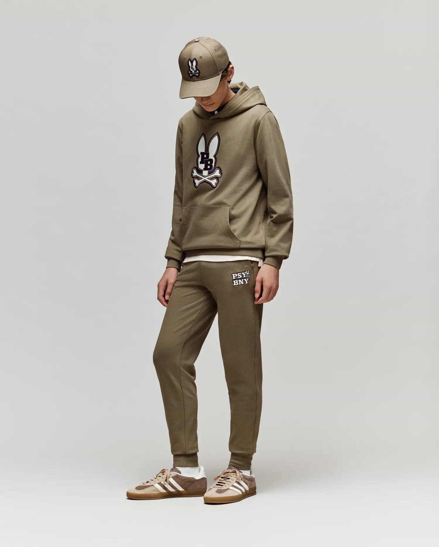 Dressed in Psycho Bunny’s KIDS WADE CORD PATCH SWEATPANT (KB0800002), a child stands against a plain light gray background, looking down with hands in pockets. They pair the brown sweatpants and hoodie with matching sneakers and a baseball cap.