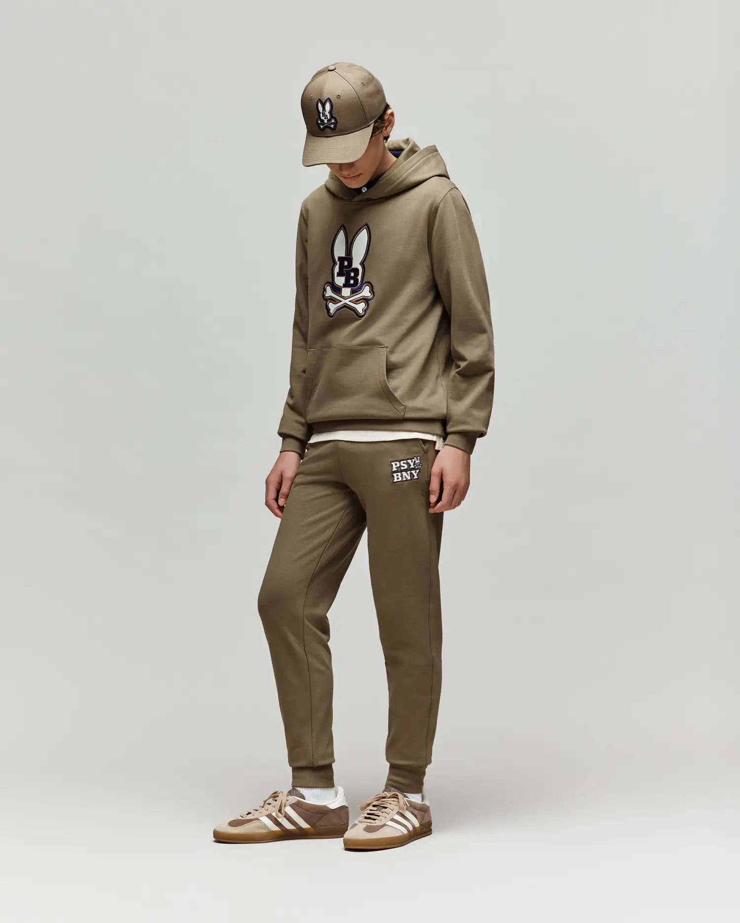 Dressed in Psycho Bunny’s KIDS WADE CORD PATCH SWEATPANT (KB0800002), a child stands against a plain light gray background, looking down with hands in pockets. They pair the brown sweatpants and hoodie with matching sneakers and a baseball cap.