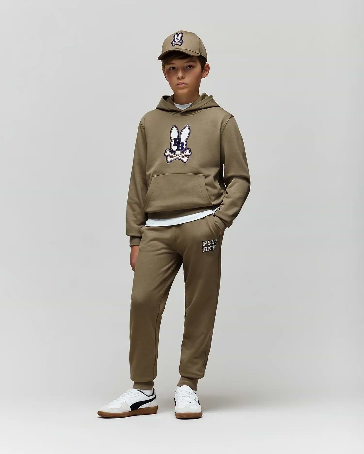 A boy wears Psycho Bunny’s KIDS WADE CORD PATCH SWEATPANT (KB0800002) with an olive green bunny hoodie, a matching cap, and white sneakers, standing against a plain light background.