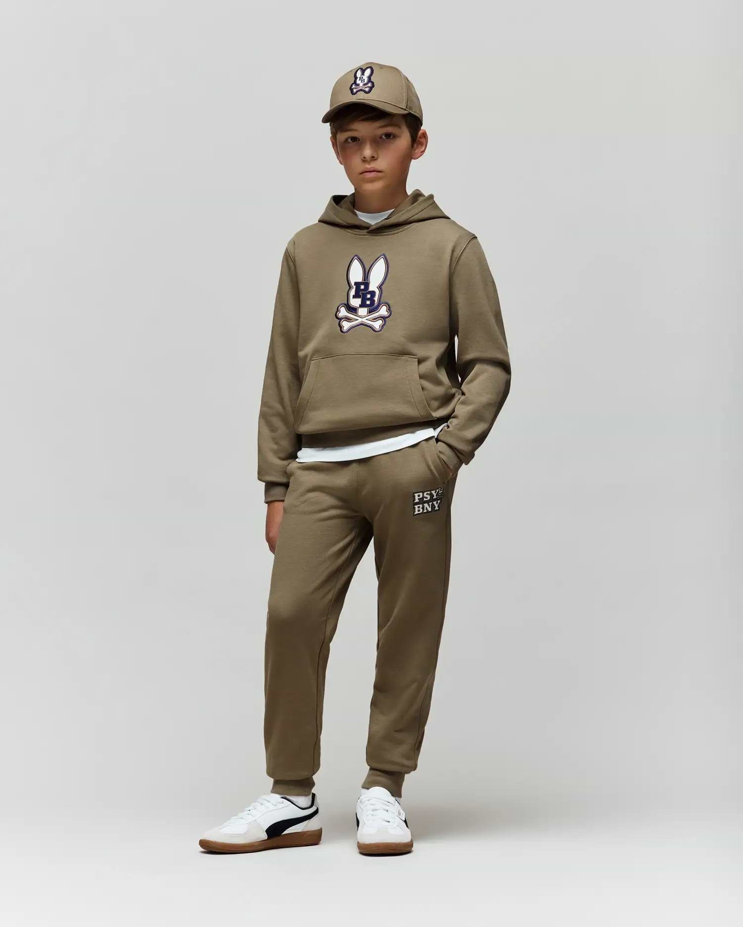 A boy wears Psycho Bunny’s KIDS WADE CORD PATCH SWEATPANT (KB0800002) with an olive green bunny hoodie, a matching cap, and white sneakers, standing against a plain light background.