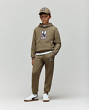 A boy wears Psycho Bunny’s KIDS WADE CORD PATCH SWEATPANT (KB0800002) with an olive green bunny hoodie, a matching cap, and white sneakers, standing against a plain light background.
