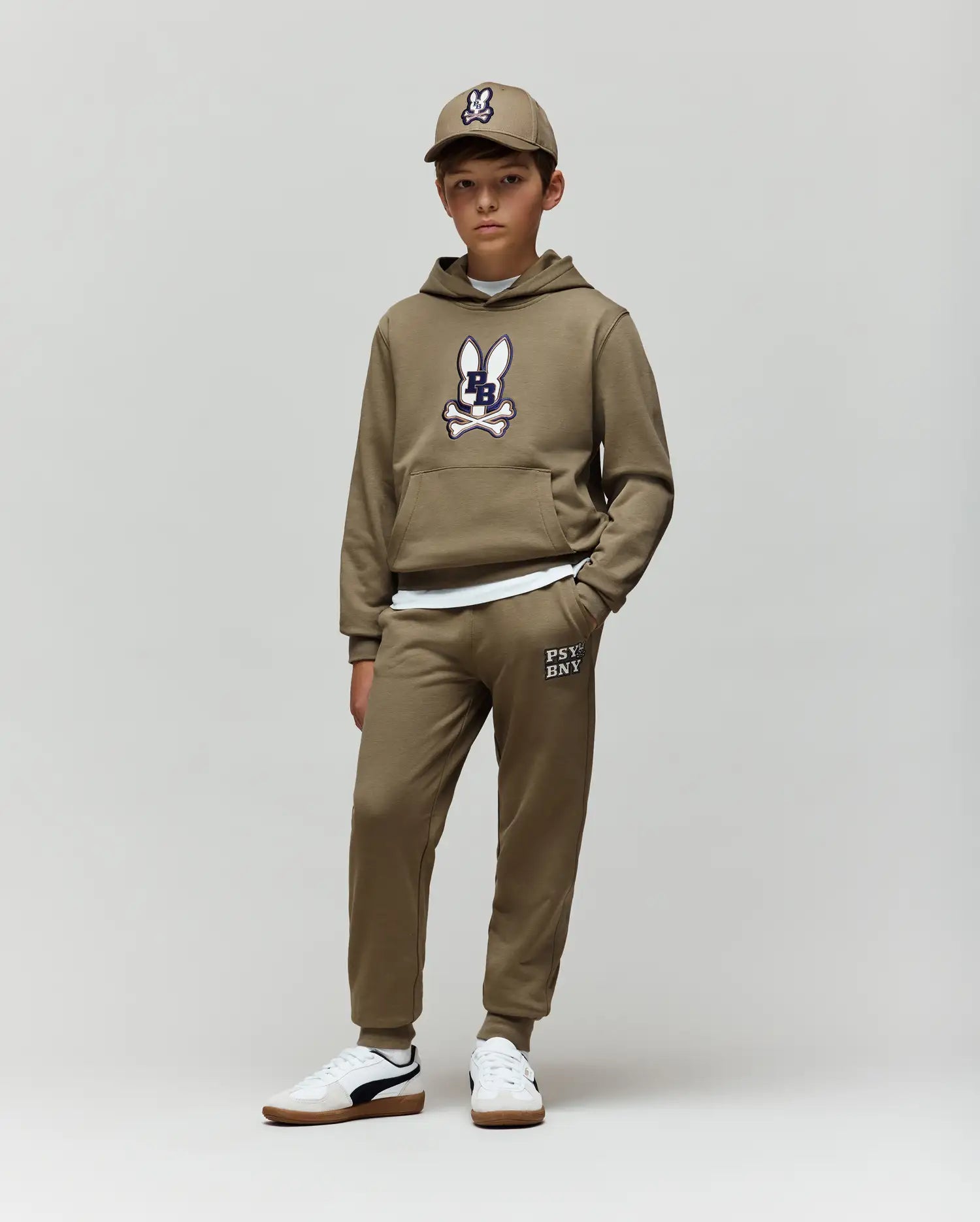 A boy wears Psycho Bunny’s KIDS WADE CORD PATCH SWEATPANT (KB0800002) with an olive green bunny hoodie, a matching cap, and white sneakers, standing against a plain light background.