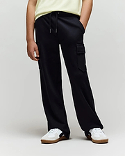 A person wearing a light yellow shirt, Psycho Bunny KIDS NASH CARGO SWEATPANT - KB0800004, and white sneakers with brown soles stands with hands in pockets against a plain background, blending street style and sweatpant comfort.