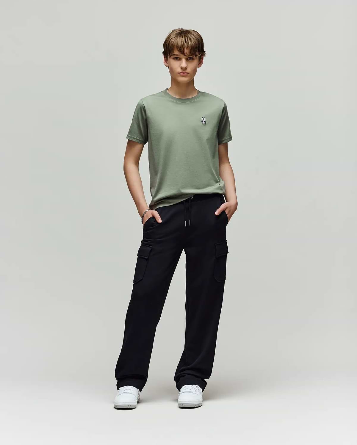A young person faces forward, wearing a light green t-shirt, white sneakers, and Psycho Bunny KIDS NASH CARGO SWEATPANT - KB0800004 with cargo pockets, hands in pockets, enjoying sweatpant comfort against a plain light gray background.