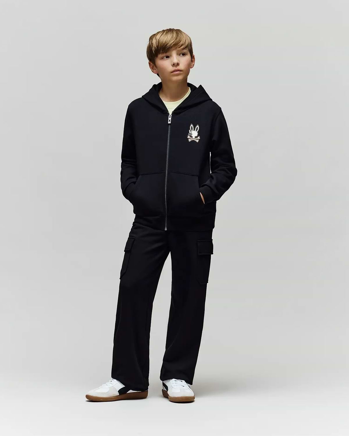 A boy stands with hands in pockets, wearing a black zip-up hoodie with a cartoon chest print, Psycho Bunny KIDS NASH CARGO SWEATPANT - KB0800004 with cargo pockets, and white sneakers, against a plain light grey background.