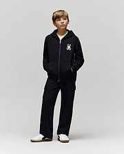 A boy stands with hands in pockets, wearing a black zip-up hoodie with a cartoon chest print, Psycho Bunny KIDS NASH CARGO SWEATPANT - KB0800004 with cargo pockets, and white sneakers, against a plain light grey background.