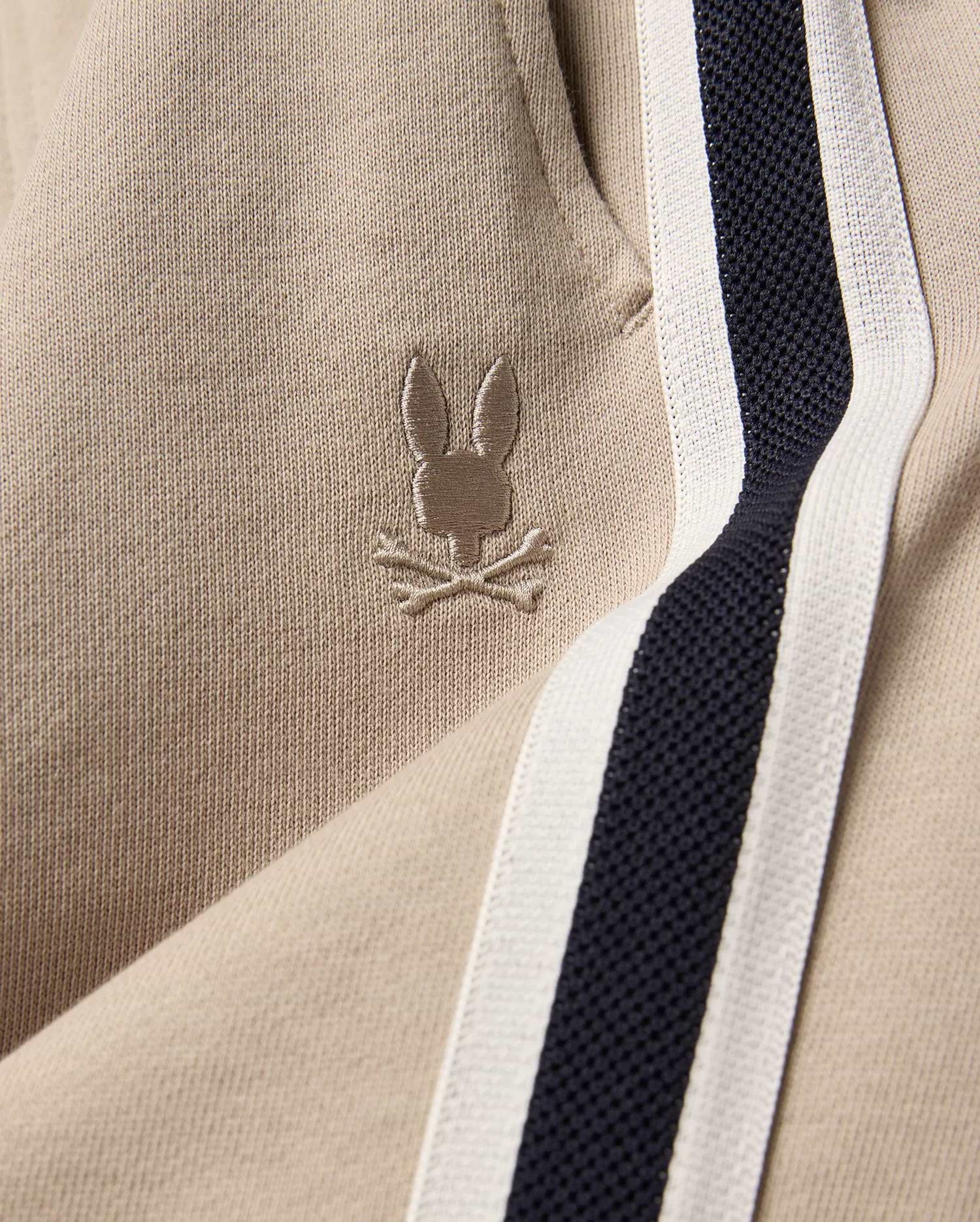 Kids Ty French Terry Sweatpant in beige french terry fabric, featuring a small embroidered Psycho Bunny skull logo and a navy and white side stripe.