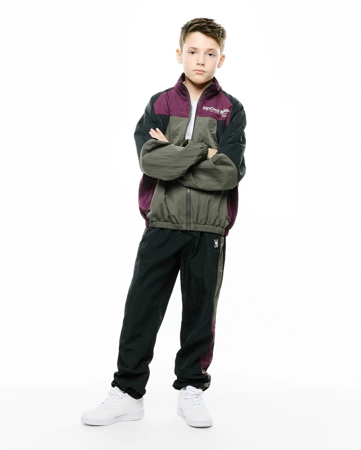 KIDS ALDER NYLON COLORBLOCK PANT in dark green/black with plum side stripe, featuring a small embroidered white Psycho Bunny skull logo on the left thigh. Relaxed fit crinkled nylon, part of a matching colorblock tracksuit. (1)
