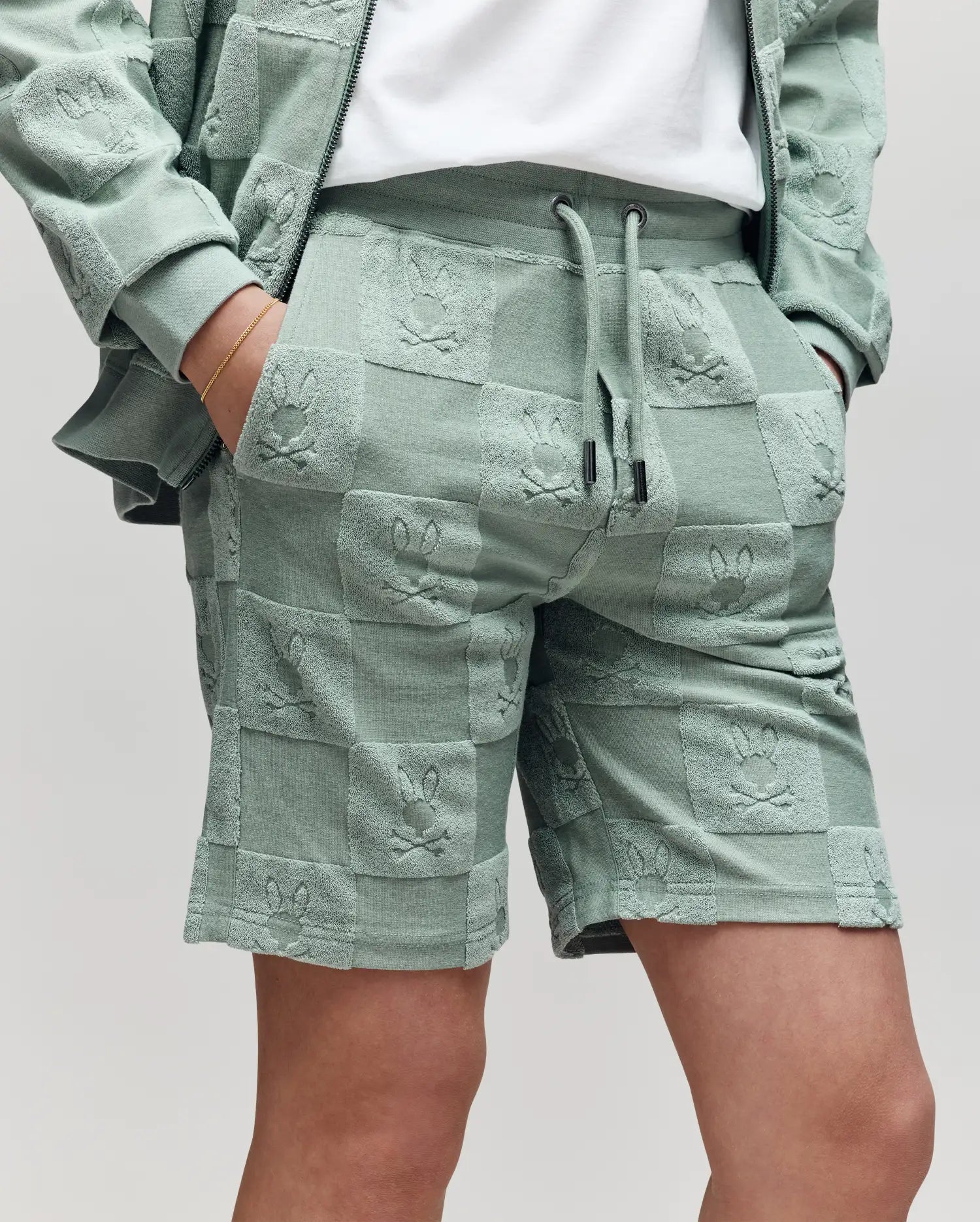 Teen | A person wears Psycho Bunny's KIDS KELVIN TOWEL TERRY SWEATSHORT (KB0900002) and a matching jacket in sage green jacquard terry, both embossed with a checkered bunny pattern featuring repeating Bugs Bunny faces, hand in pocket.