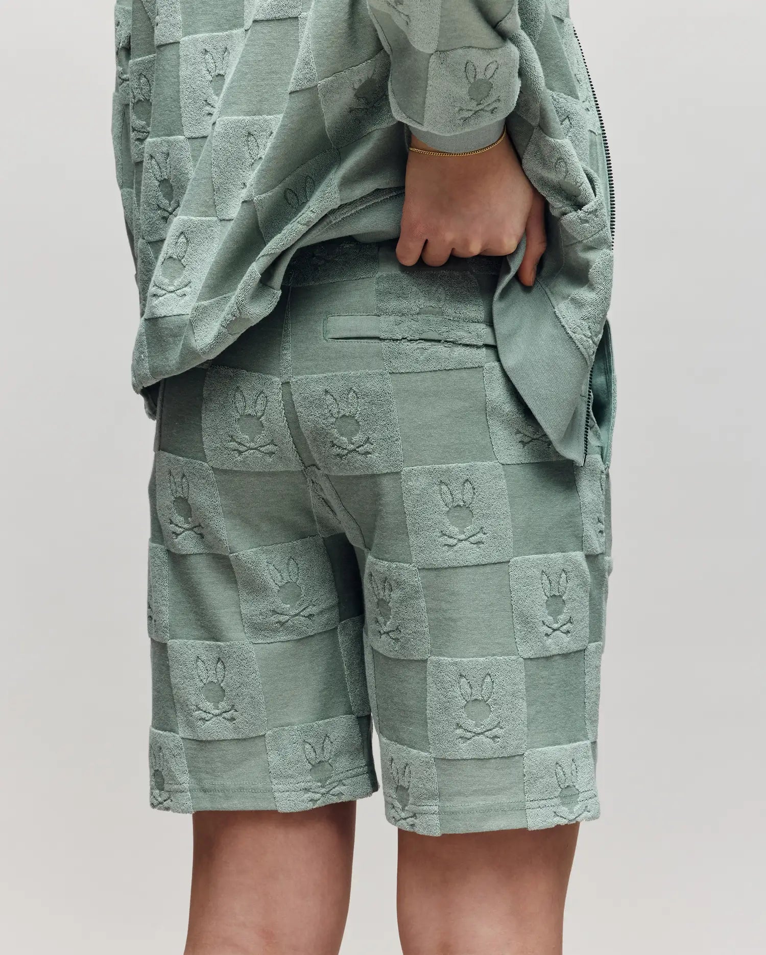 Teen | A child models Psycho Bunny's KIDS KELVIN TOWEL TERRY SWEATSHORT (KB0900002) in textured sage green cotton blend, paired with a matching checkered bunny jacket. The back view shows one hand resting on their hip.