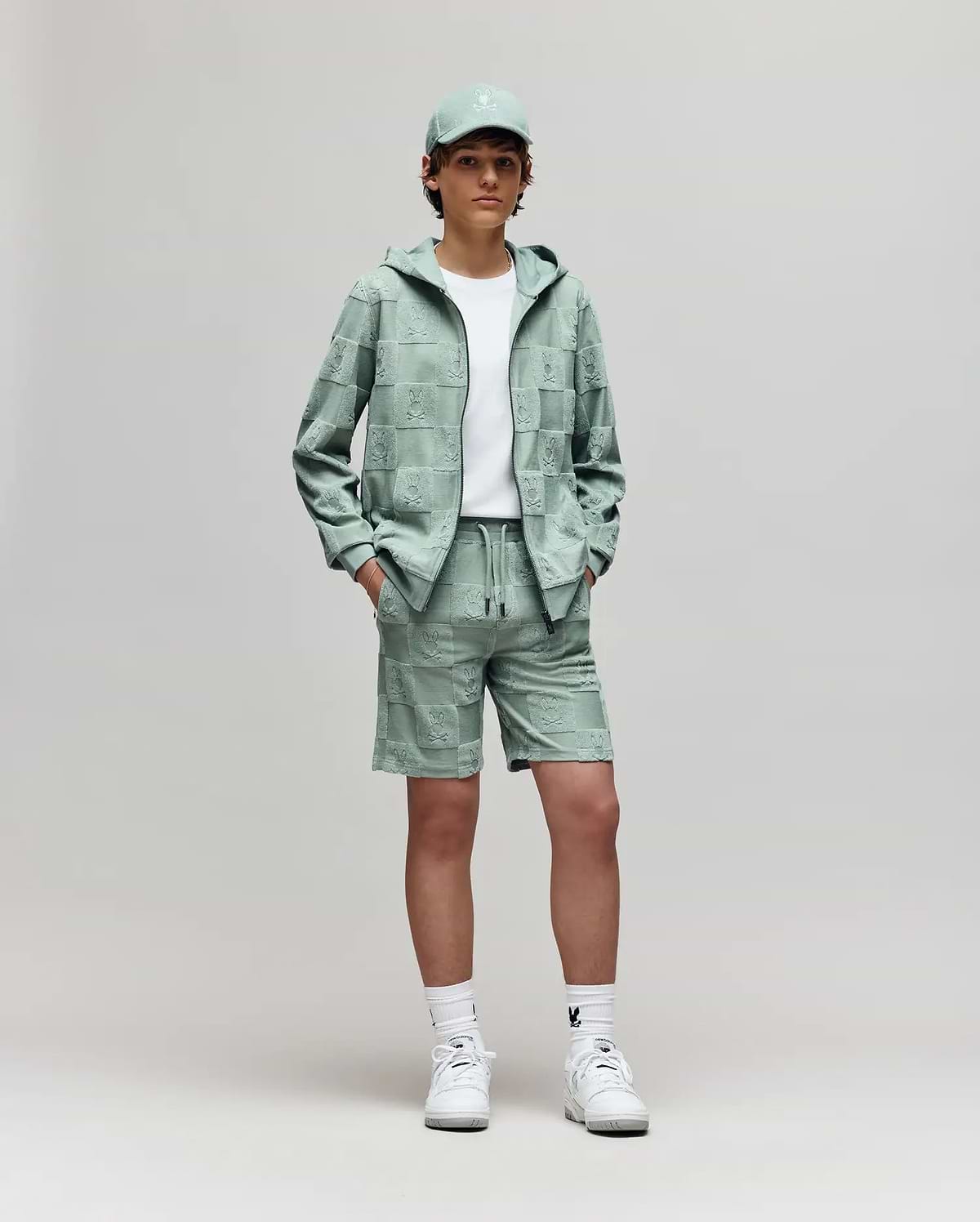Teen | A young person stands against a plain background, wearing Psycho Bunny's KIDS KELVIN TOWEL TERRY SWEATSHORT (KB0900002) with a white tee, mint green cap and hoodie, and white sneakers, hands in pockets, looking at the camera.