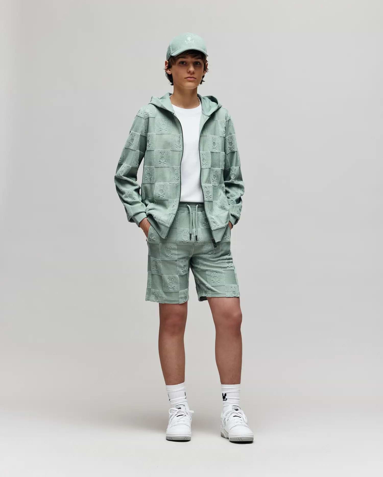 Teen | A young person stands against a plain background, wearing Psycho Bunny's KIDS KELVIN TOWEL TERRY SWEATSHORT (KB0900002) with a white tee, mint green cap and hoodie, and white sneakers, hands in pockets, looking at the camera.