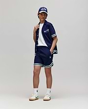 Teen | A person poses against a plain background wearing Psycho Bunny’s KIDS BASIL TAPED SHORT - KB0900003 with white and green trim, a white cotton-modal T-shirt, white sneakers, and a navy cap. Their hand rests on their collar as they face the camera.