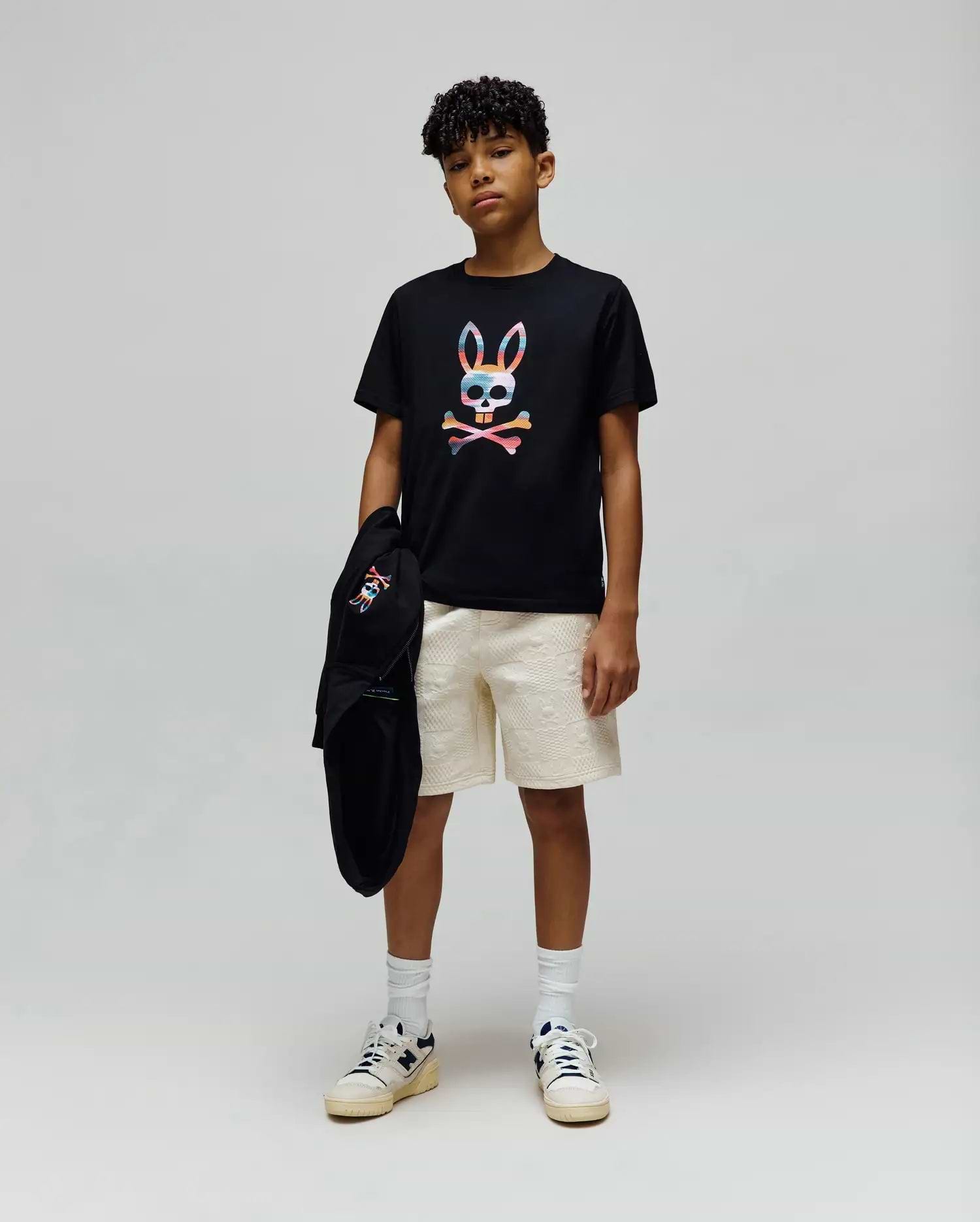 Black jersey t-shirt with a large colorful Psycho Bunny skull graphic, paired with cream Kids Dustin Sweatshort featuring a subtle textured pattern, and a black hoodie with a small embroidered bunny logo.