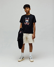 Black jersey t-shirt with a large colorful Psycho Bunny skull graphic, paired with cream Kids Dustin Sweatshort featuring a subtle textured pattern, and a black hoodie with a small embroidered bunny logo.