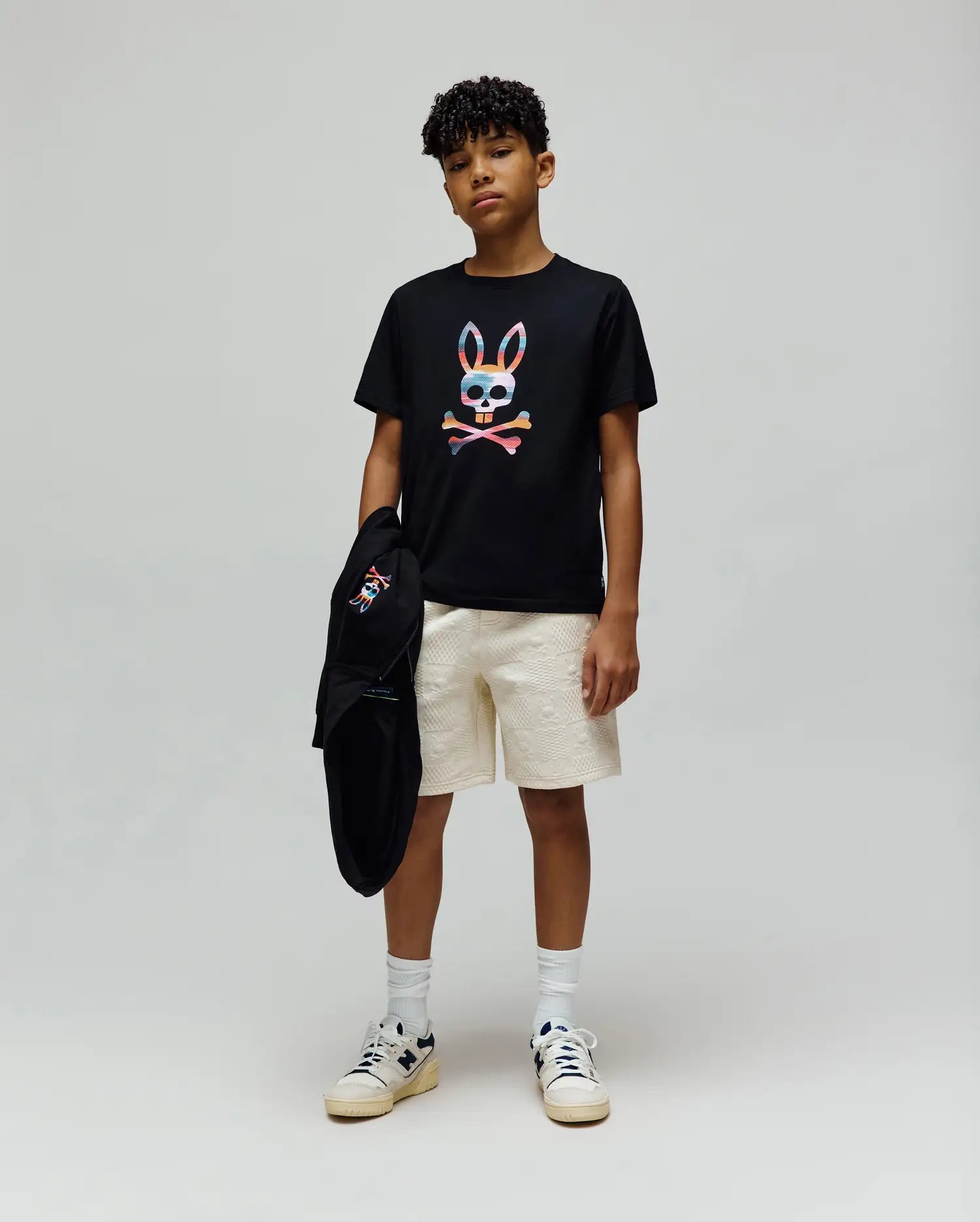 Black jersey t-shirt with a large colorful Psycho Bunny skull graphic, paired with cream Kids Dustin Sweatshort featuring a subtle textured pattern, and a black hoodie with a small embroidered bunny logo.