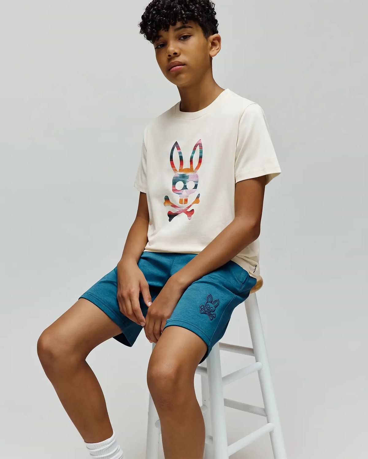 Cream jersey t-shirt with a large multi-color plaid Psycho Bunny logo on the chest, paired with Kids Barry Modal Short in teal, featuring a small embroidered bunny skull logo on the left thigh.