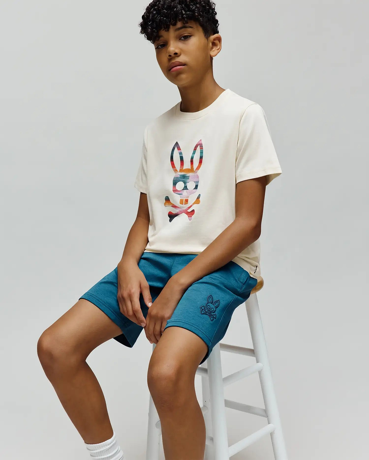 Cream jersey t-shirt with a large multi-color plaid Psycho Bunny logo on the chest, paired with Kids Barry Modal Short in teal, featuring a small embroidered bunny skull logo on the left thigh.