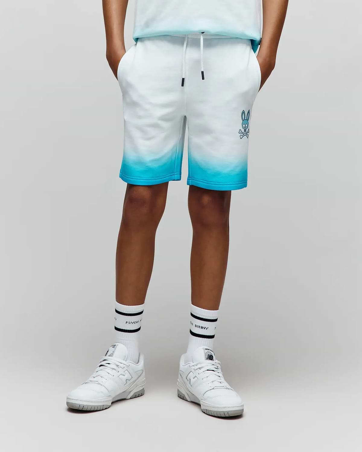 Kids Dax Sweatshort in white french terry, featuring a vibrant blue ombre fade from mid-thigh to hem, with an embroidered blue Psycho Bunny skull logo on the left thigh and a drawstring waist.